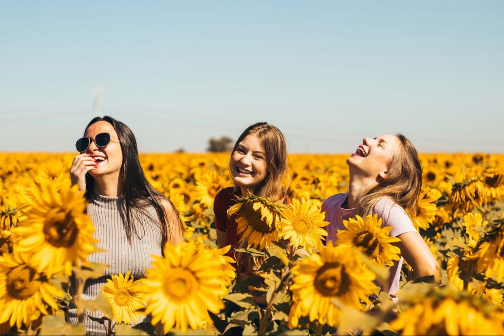 Three women laughing joyfully in a sunflower field on a sunny day.